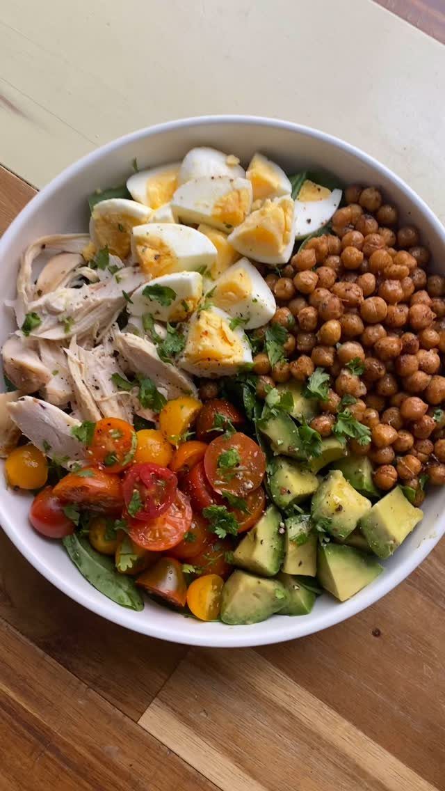 Healthy bowl with eggs, chicken, chickpeas, tomatoes, and avocado on a wooden table.