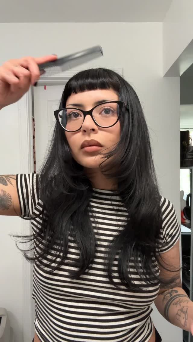 Woman with long black hair and blunt bangs wearing black glasses and striped shirt.