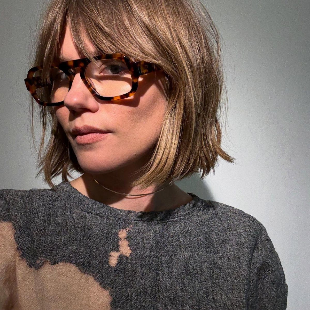Woman with blonde bob and bangs wearing tortoiseshell glasses against a grey wall.