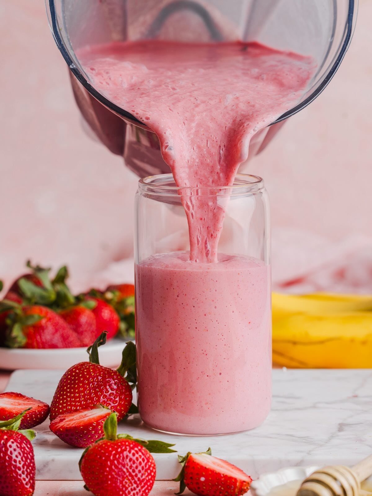 Pink strawberry smoothie pouring into a glass with fresh strawberries and bananas nearby.