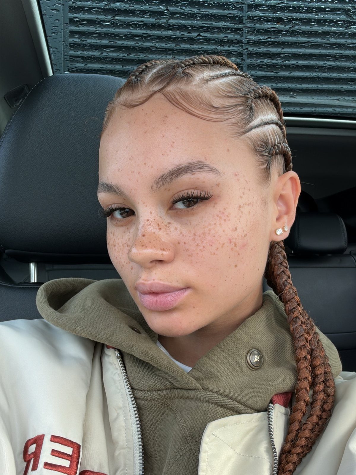 Person with brown feed-in cornrow braids wearing a green hoodie in a car.