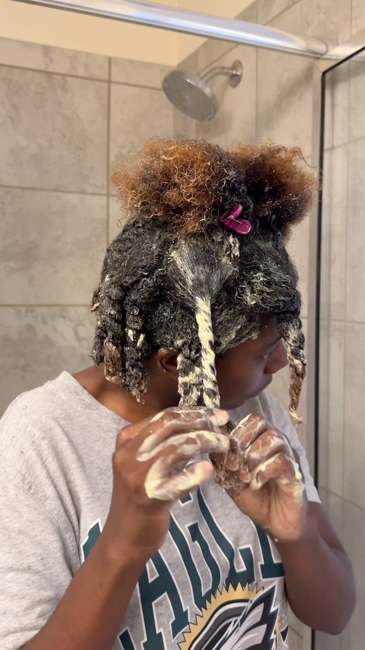 Person applying a thick creamy hair mask to curly hair in a bathroom.