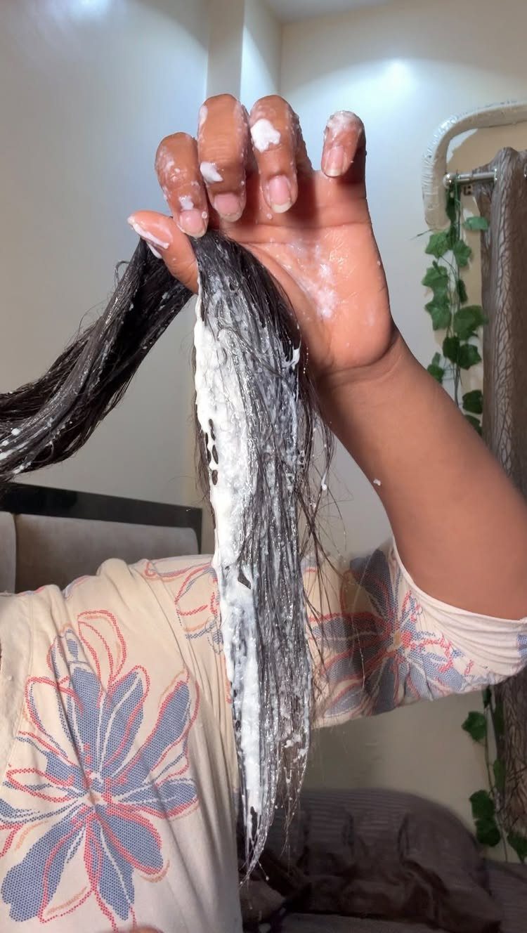 Hand applying a white creamy hair mask to a strand of long dark hair.