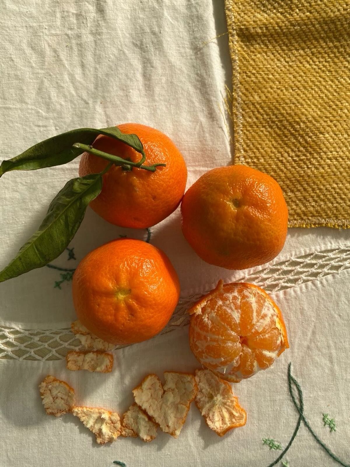 Four orange tangerines on a white cloth, one partially peeled with green leaves.