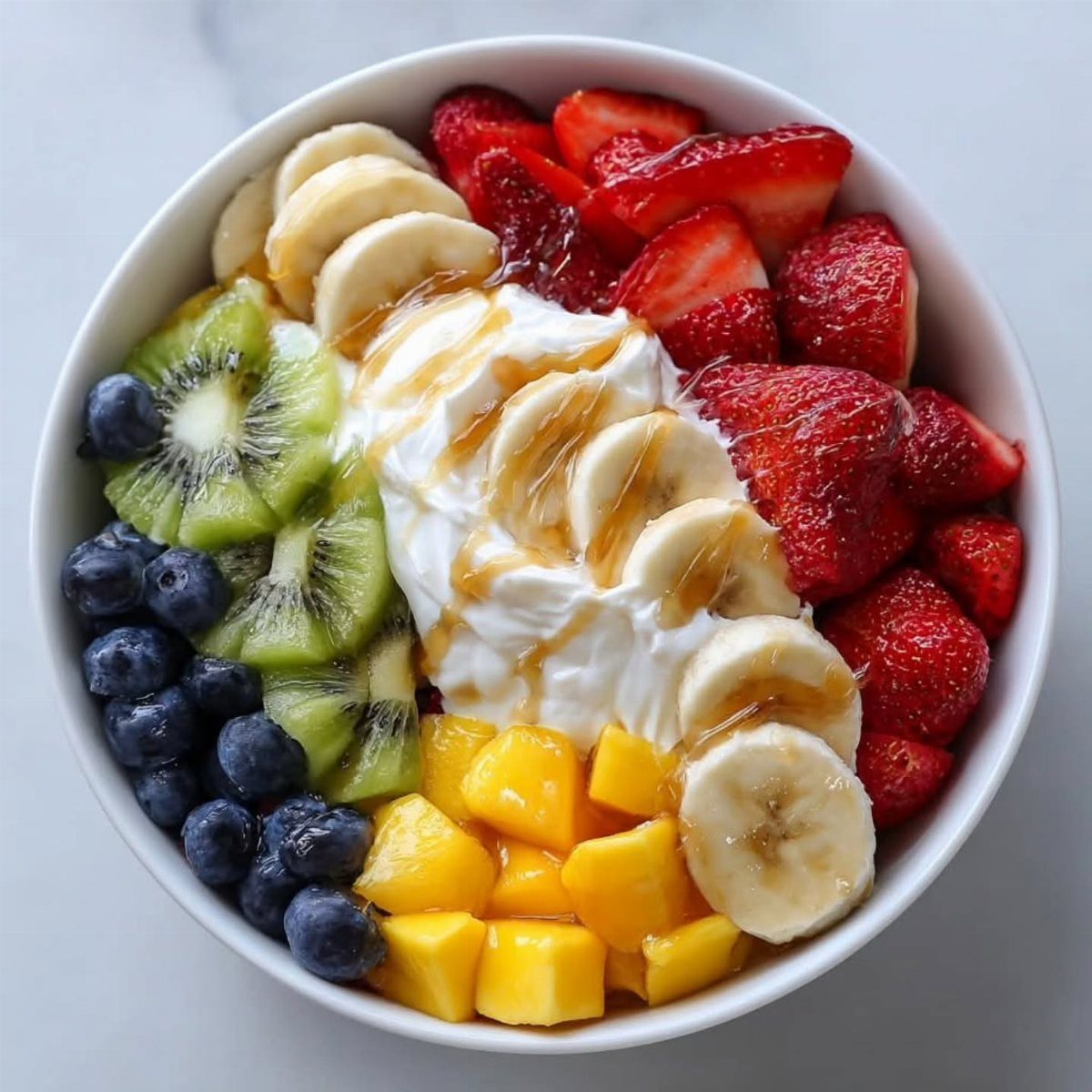 Fruit bowl with strawberries, bananas, kiwi, blueberries, mango, and yogurt with honey drizzle.