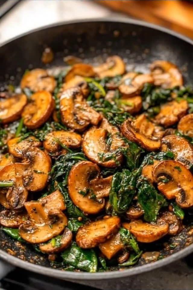 Sautéed brown mushrooms and green spinach leaves in a dark grey frying pan.