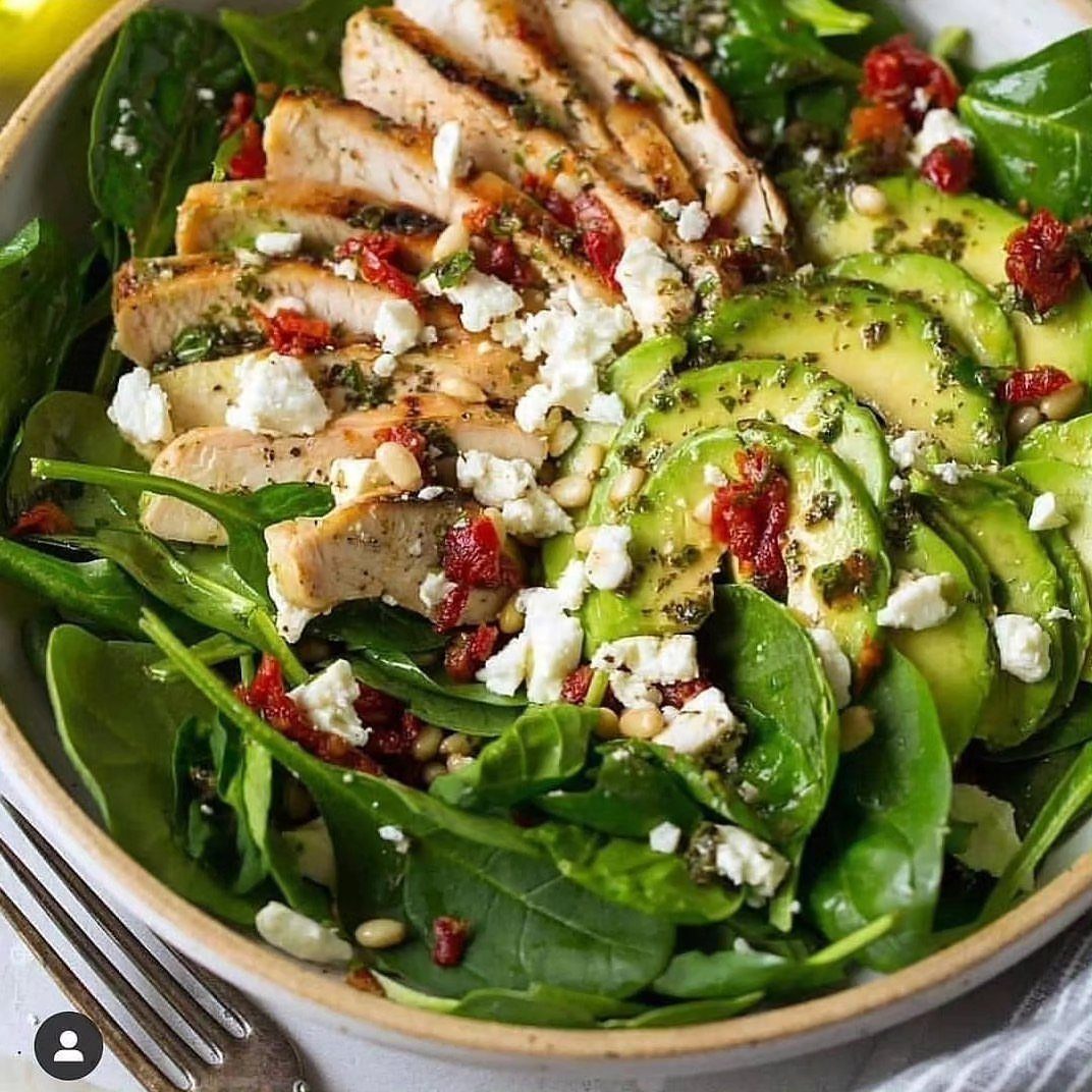 Spinach salad with grilled chicken, avocado, feta cheese, and sun-dried tomatoes in a bowl.