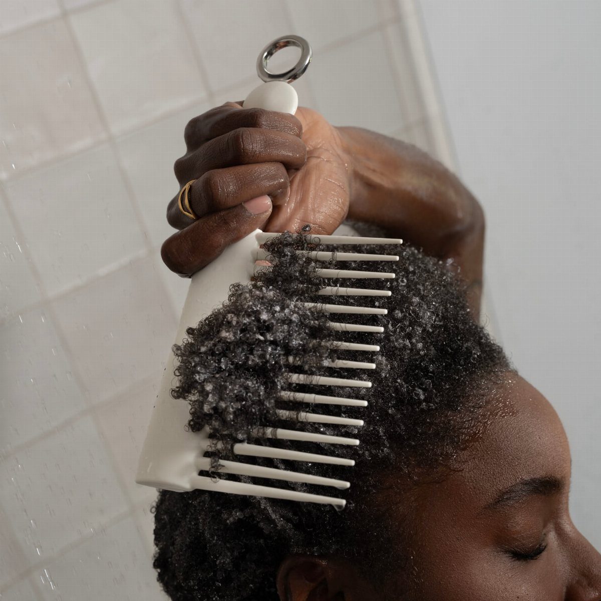 Person with curly hair using a white wide-tooth comb in a white tiled shower.