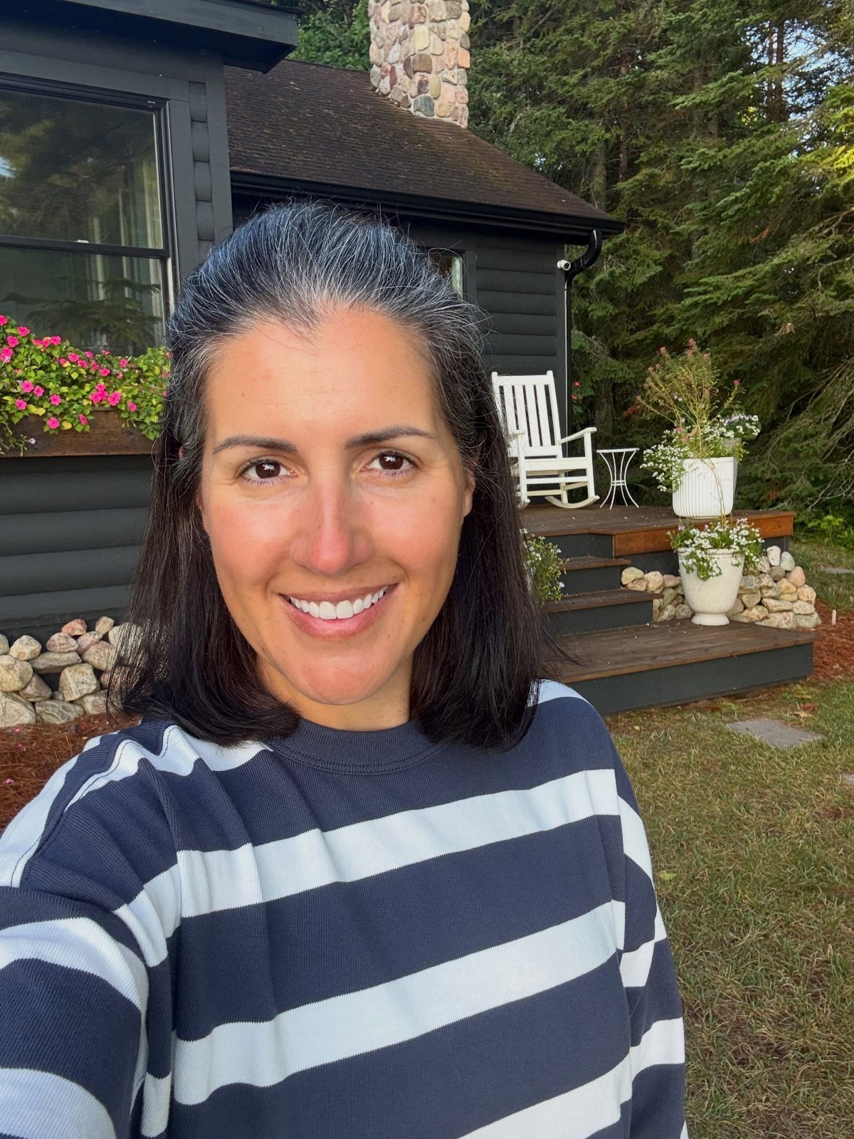 Woman with dark hair and gray roots smiling outdoors by a house.