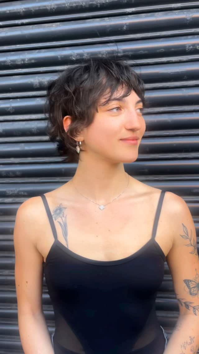 Woman with dark curly grow-out pixie hair posing against a grey metal shutter.