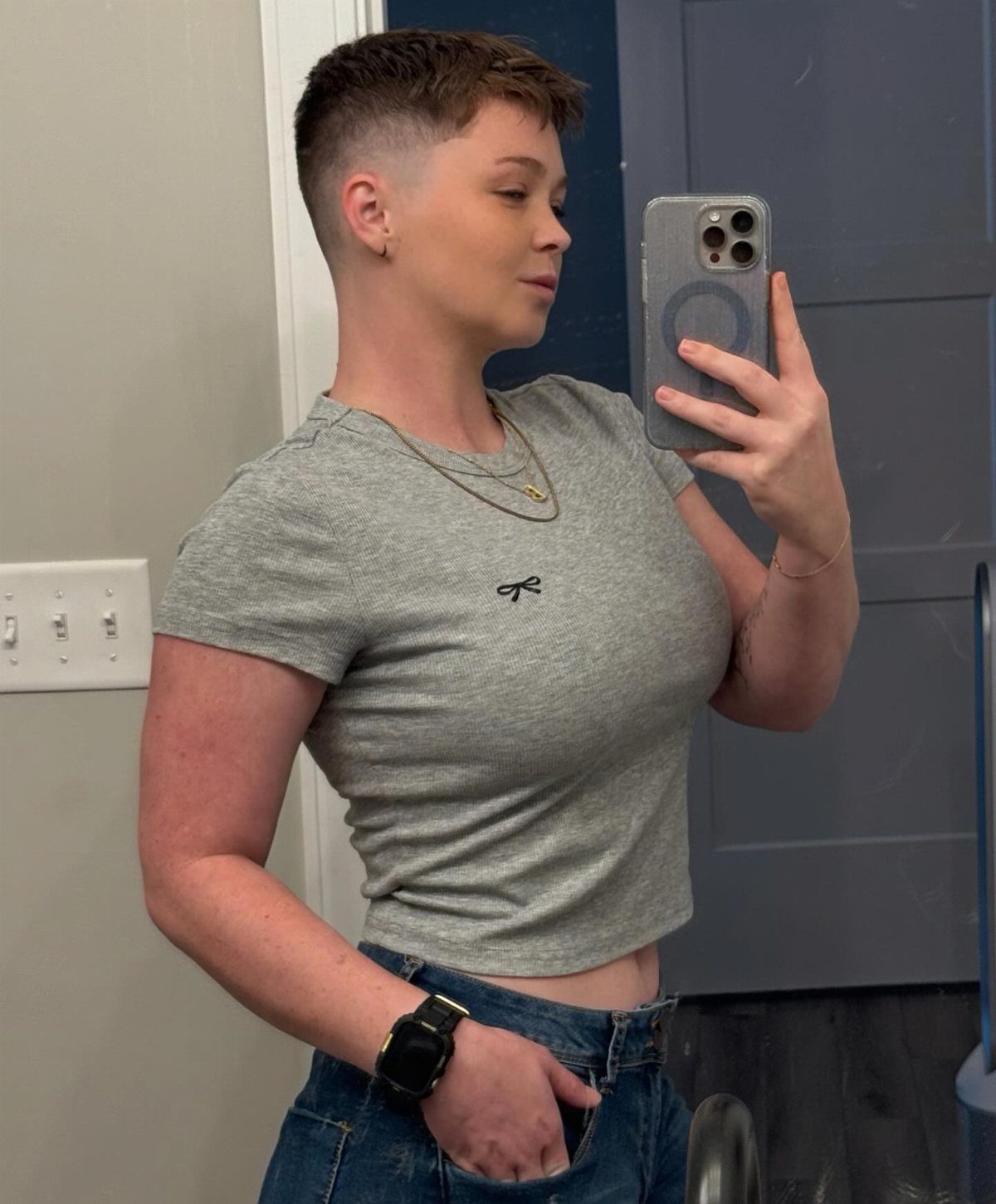 Person with a tapered pixie haircut taking a mirror selfie in a grey shirt.