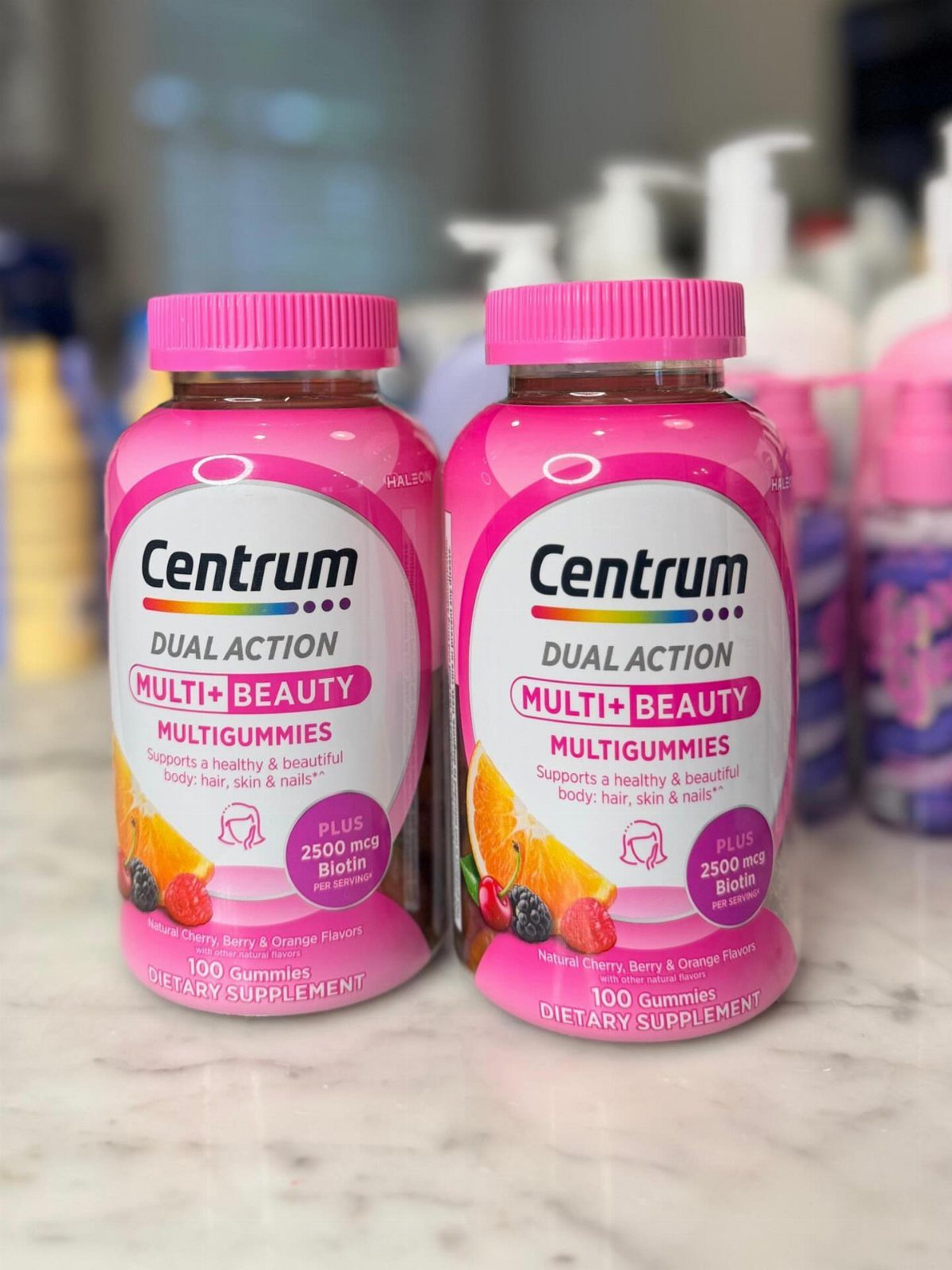 Two pink bottles of Centrum Multi+Beauty Multigummies on a white marble countertop.