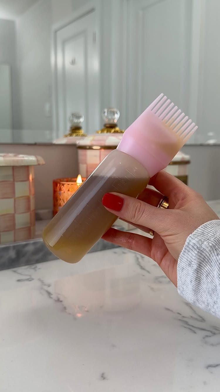Hand holding a bottle of brown hair oil with a pink comb applicator.
