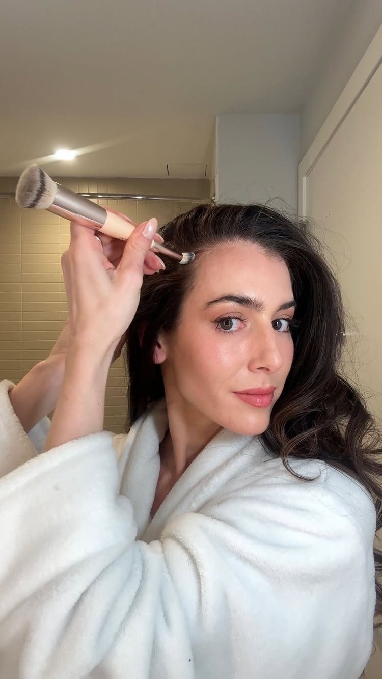 Woman in white bathrobe applying product to her hairline with a makeup brush.