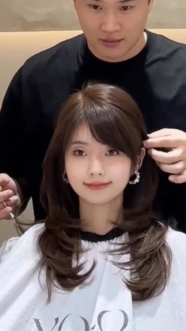Woman with long brown hair and side-swept bangs being styled by a hairdresser.