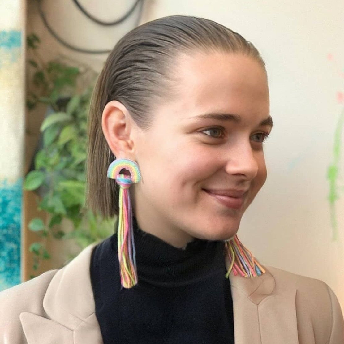 Woman with brown hair slicked back wearing rainbow tassel earrings and a beige blazer.