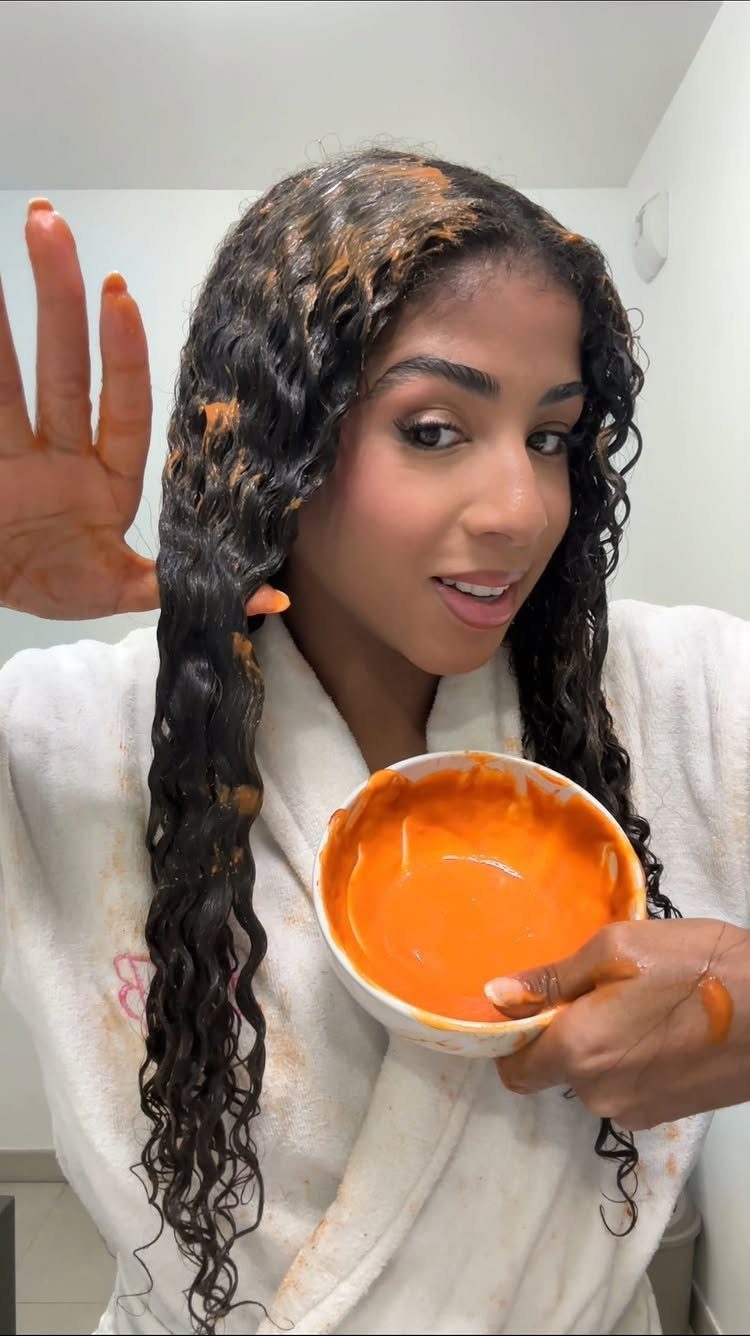 Woman with curly hair applying an orange hair mask from a white bowl.