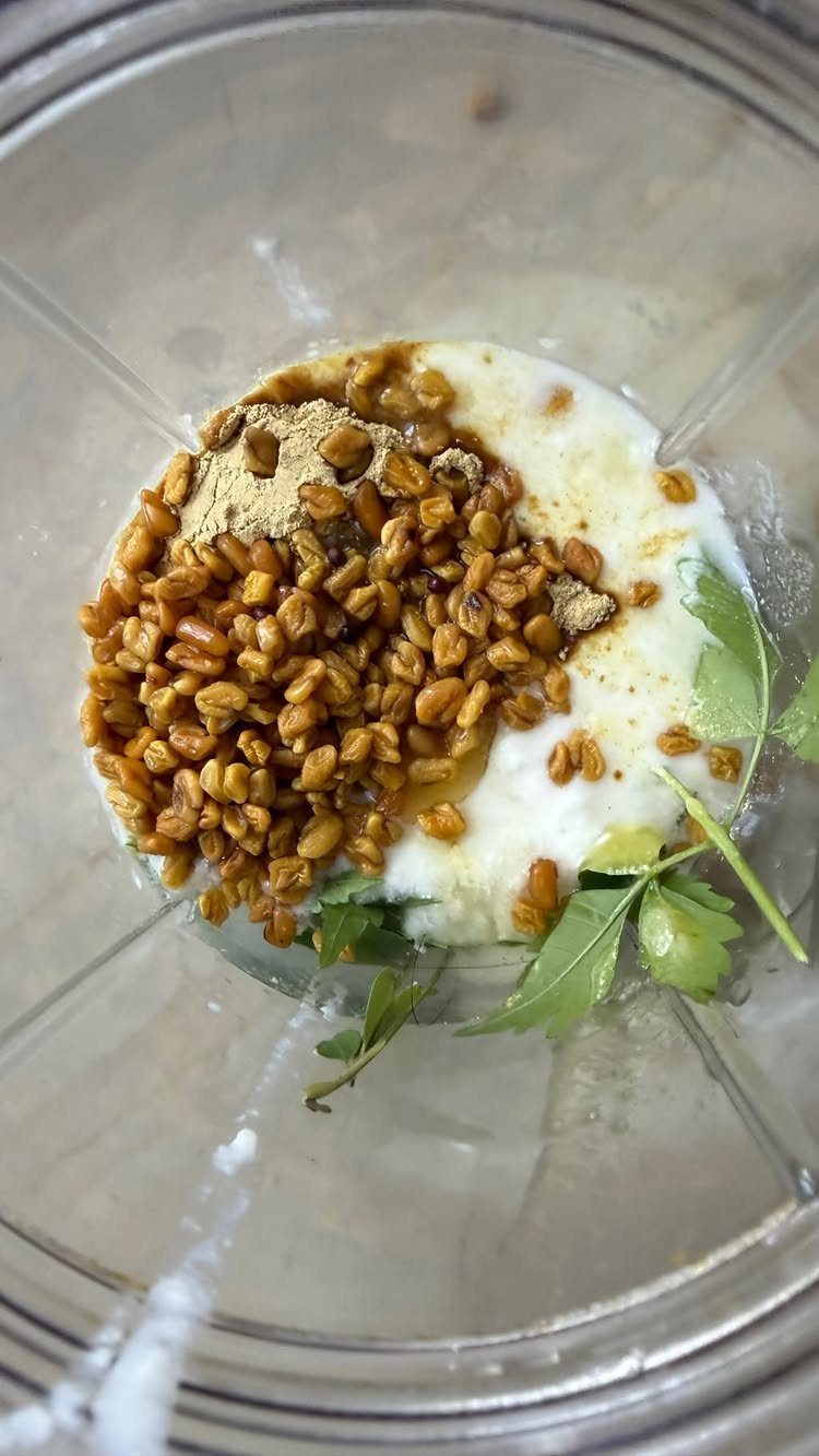 Yogurt, brown seeds, powder, and green leaves inside a clear blender jar.