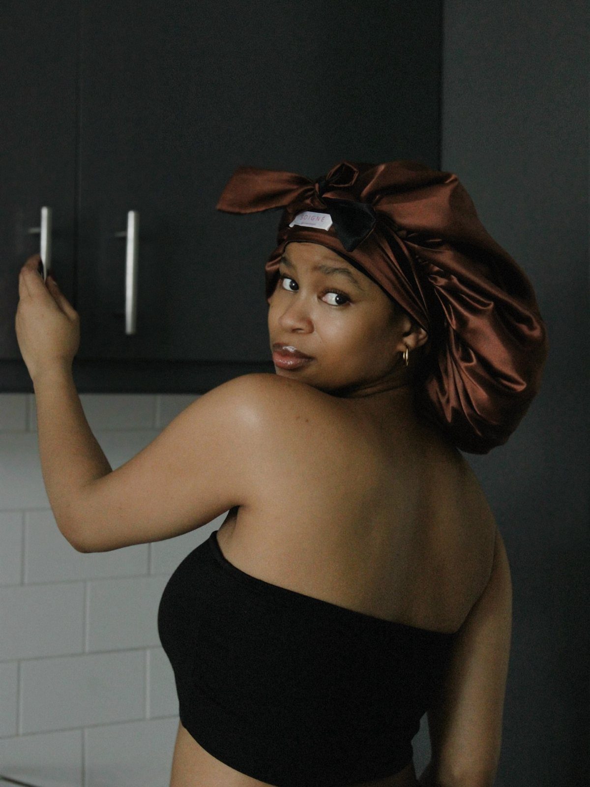 Woman wearing a brown satin bonnet and black top looking over her shoulder.