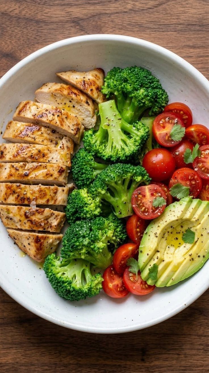Grilled chicken, broccoli, cherry tomatoes, and avocado in a white bowl on wood.