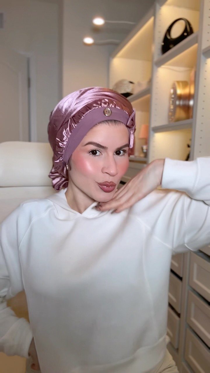 Woman wearing a pink satin protective bonnet and white sweatshirt in a bright room.