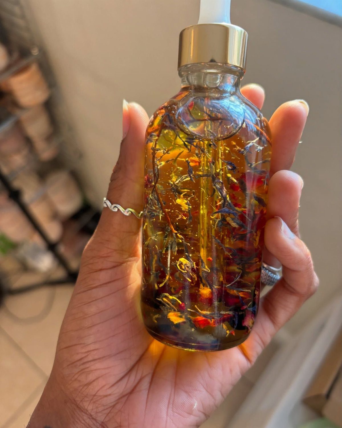 Hand holding a glass bottle of golden hair oil infused with dried botanicals.