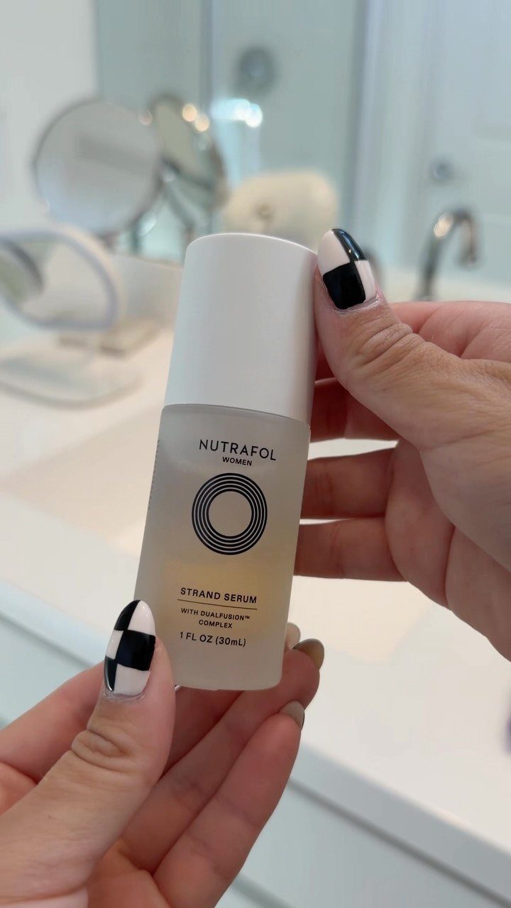 Nutrafol Strand Serum bottle held by hands in a bright white bathroom setting.