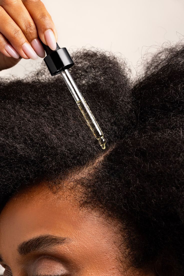Glass dropper applying clear hair growth oil to a person's dark curly scalp