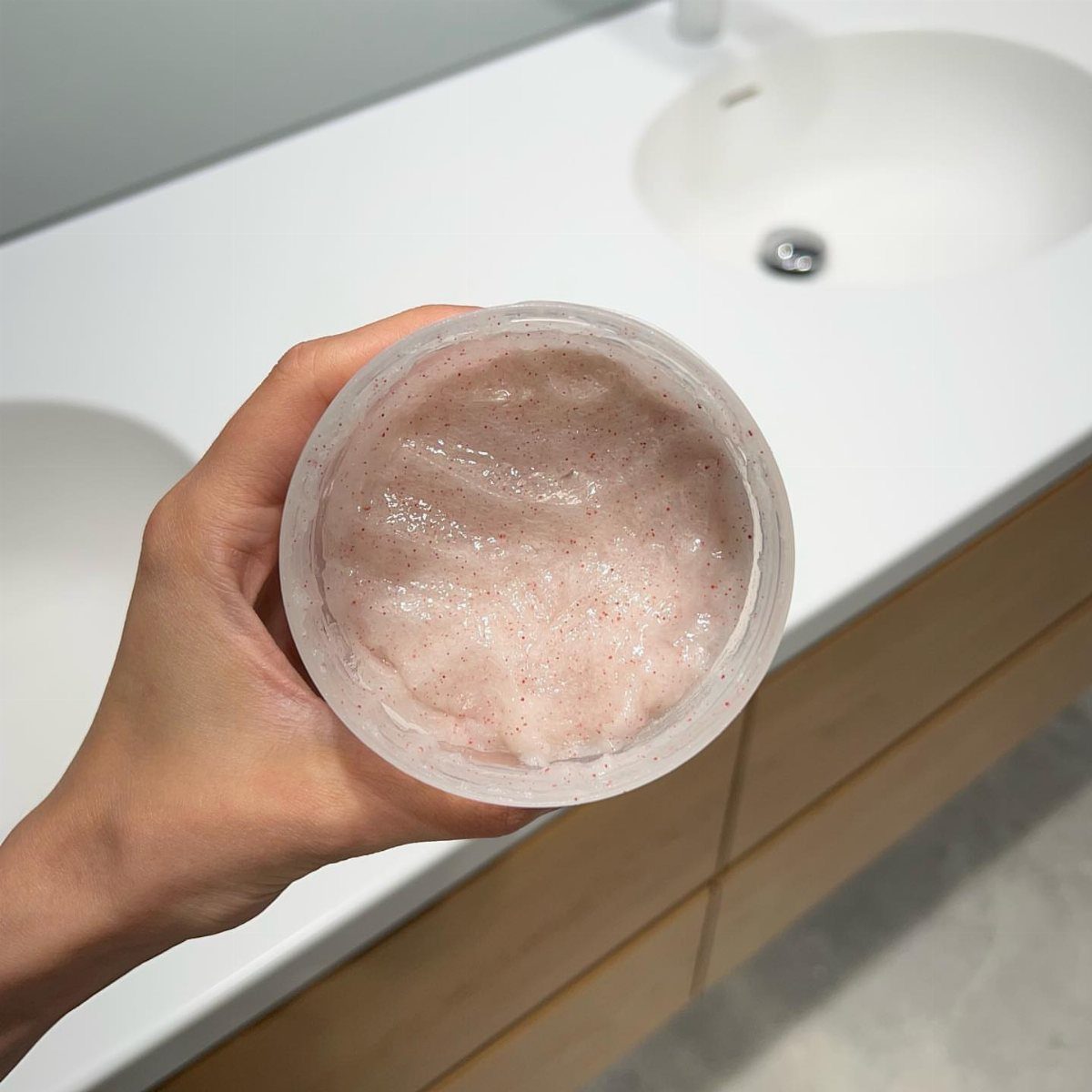 Hand holding a clear jar of grainy pink salt scalp scrub in a bathroom.