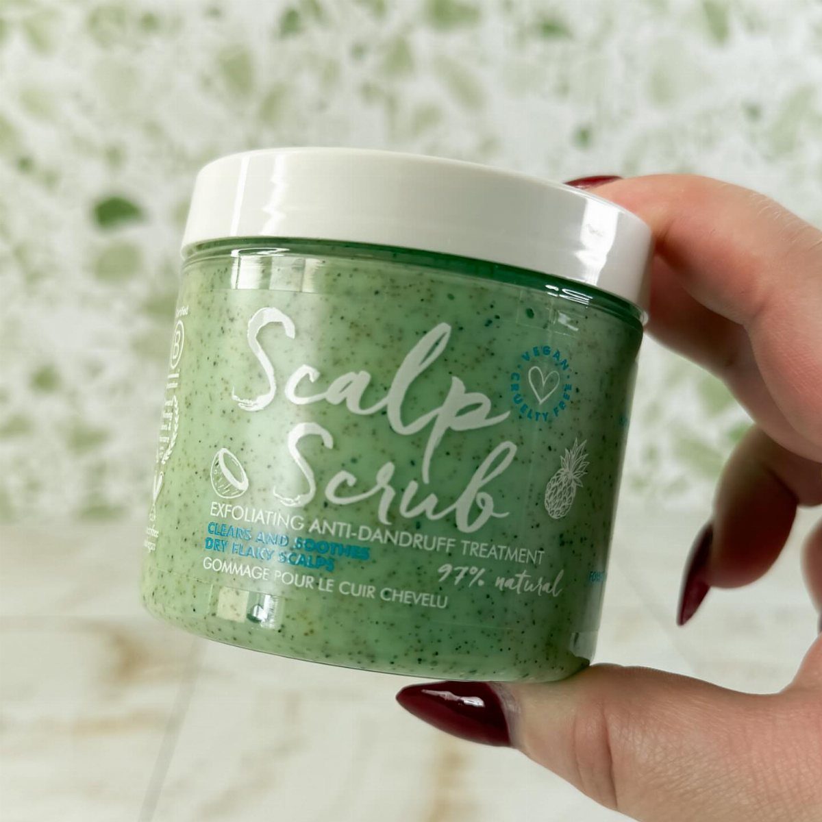 Green grainy scalp scrub in a clear jar held by a hand.