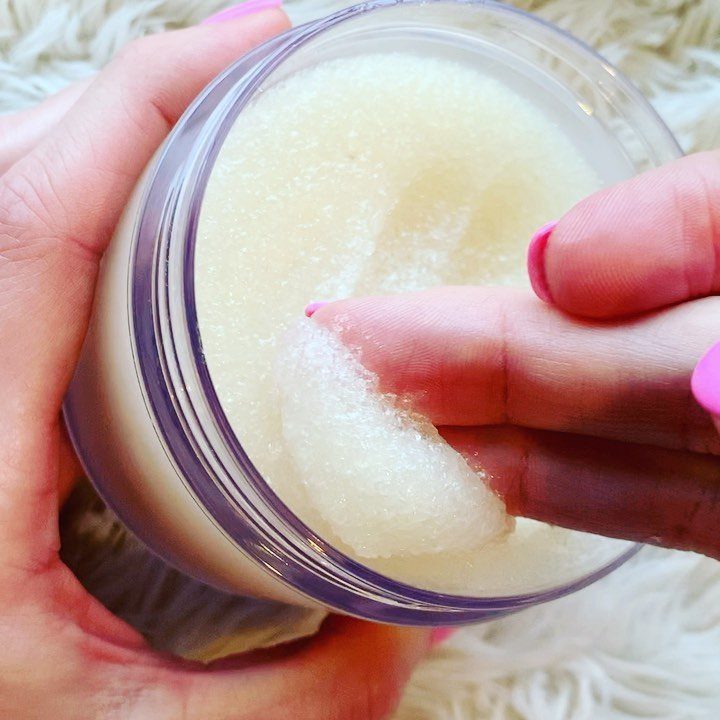 White granular salt scrub in a clear plastic jar being touched by a finger.