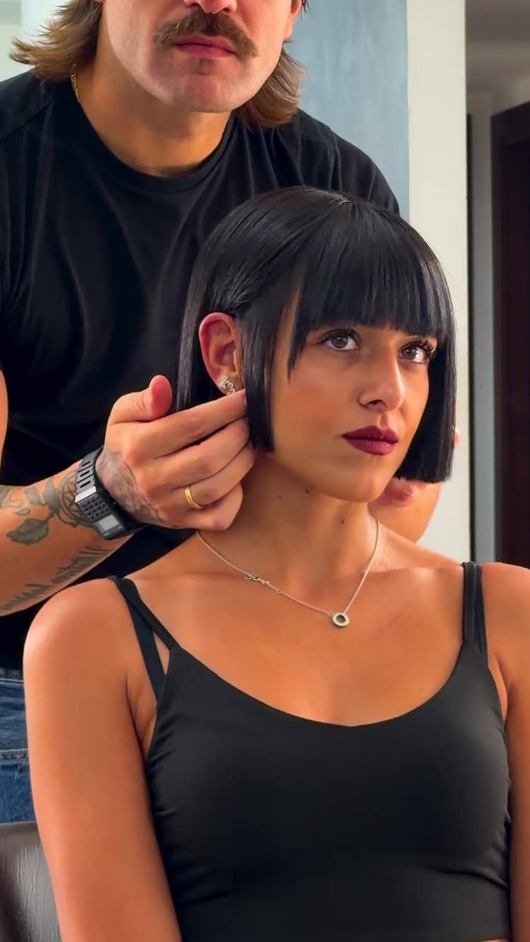Woman with a sharp black blunt bob and bangs being styled by a hairdresser.