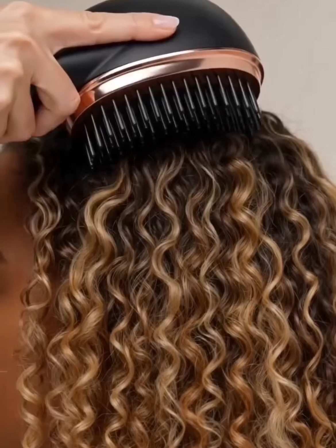Black and rose-gold hairbrush brushing through curly blonde and brown hair.