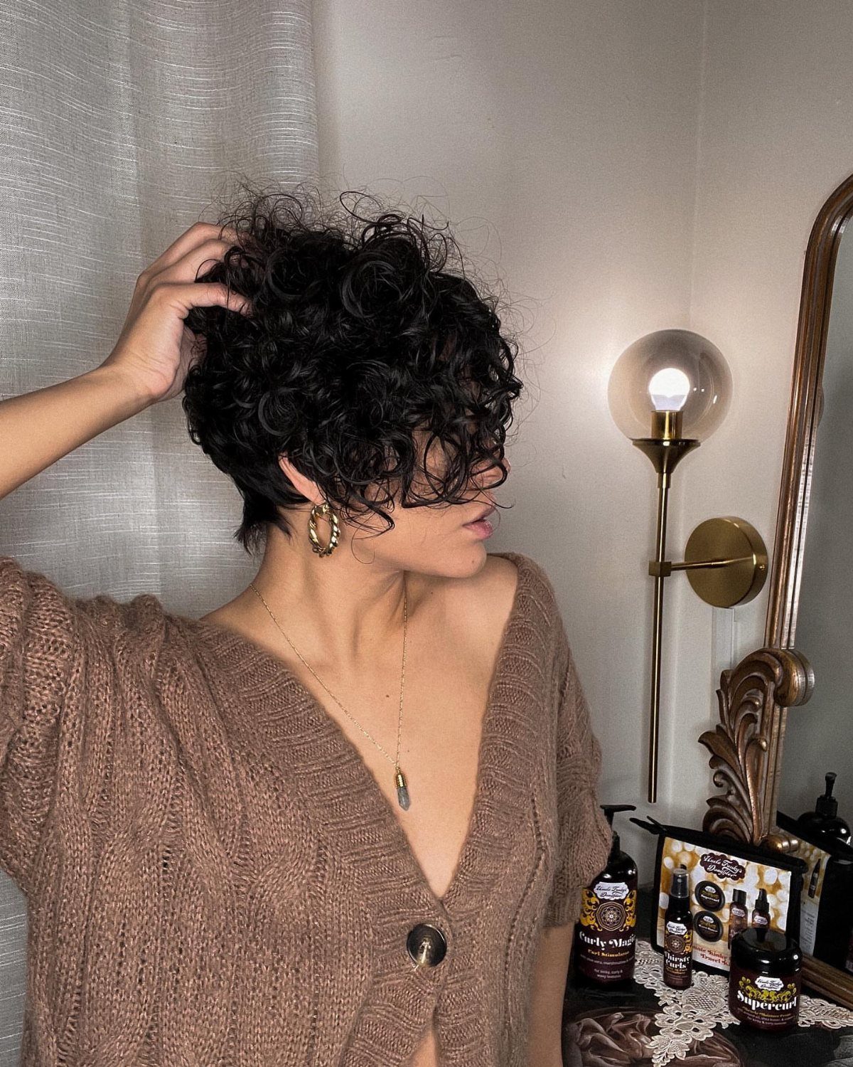 Woman with a voluminous curly pixie haircut wearing a brown knit cardigan.