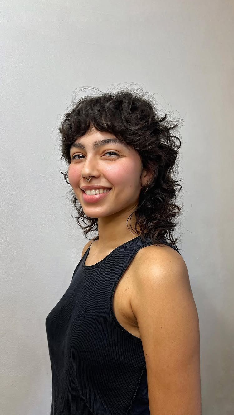 Woman with a dark wavy shag haircut and bangs smiling against a grey wall.