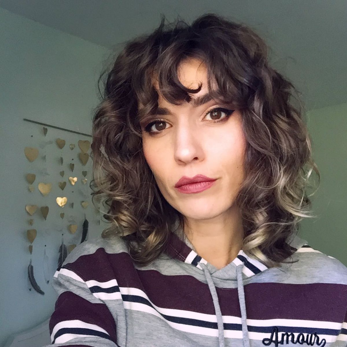 Woman with curly brown hair, wispy bangs, and blonde highlights wearing a striped hoodie.