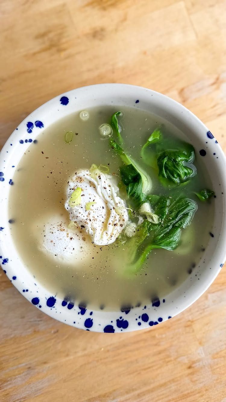 Bowl of clear broth soup with poached eggs and green leafy vegetables on wood.